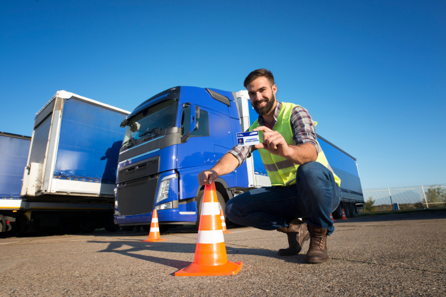 A man squatting next to a traffic cone, while holding up a CDL license in front of a blue truck.